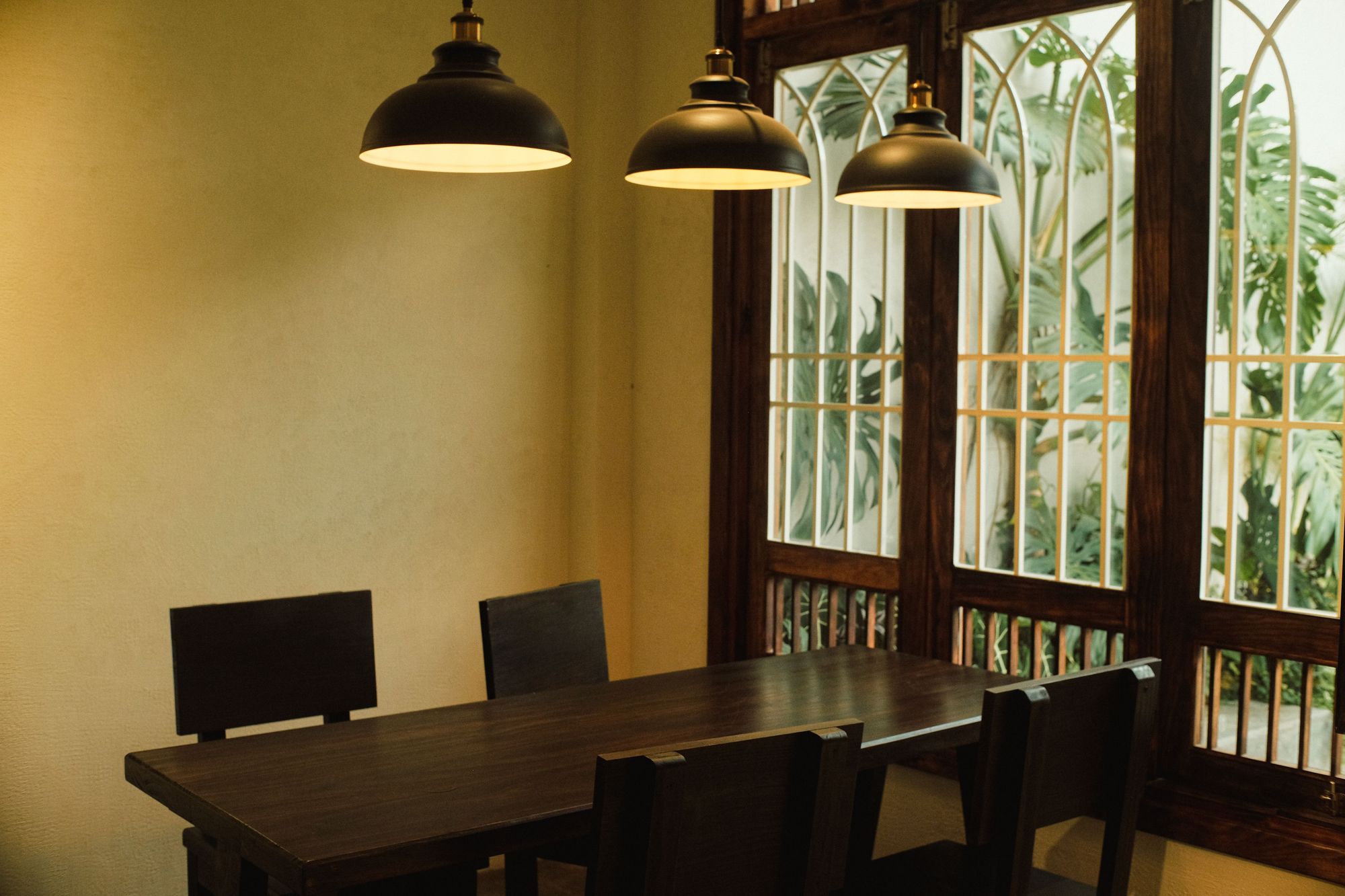 Dining room with a dark wood table, black pendant lights and tall windows framing tropical leaves