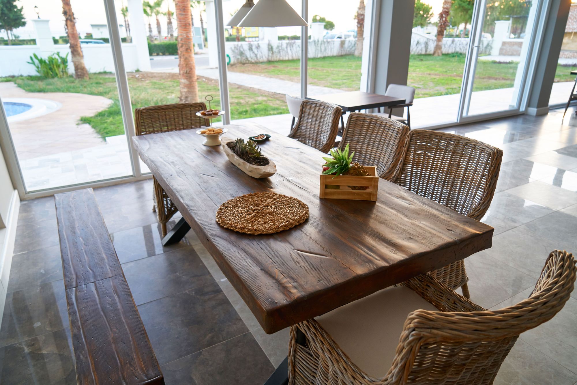 Live-edge dining table with woven chairs beside a glass wall and pool