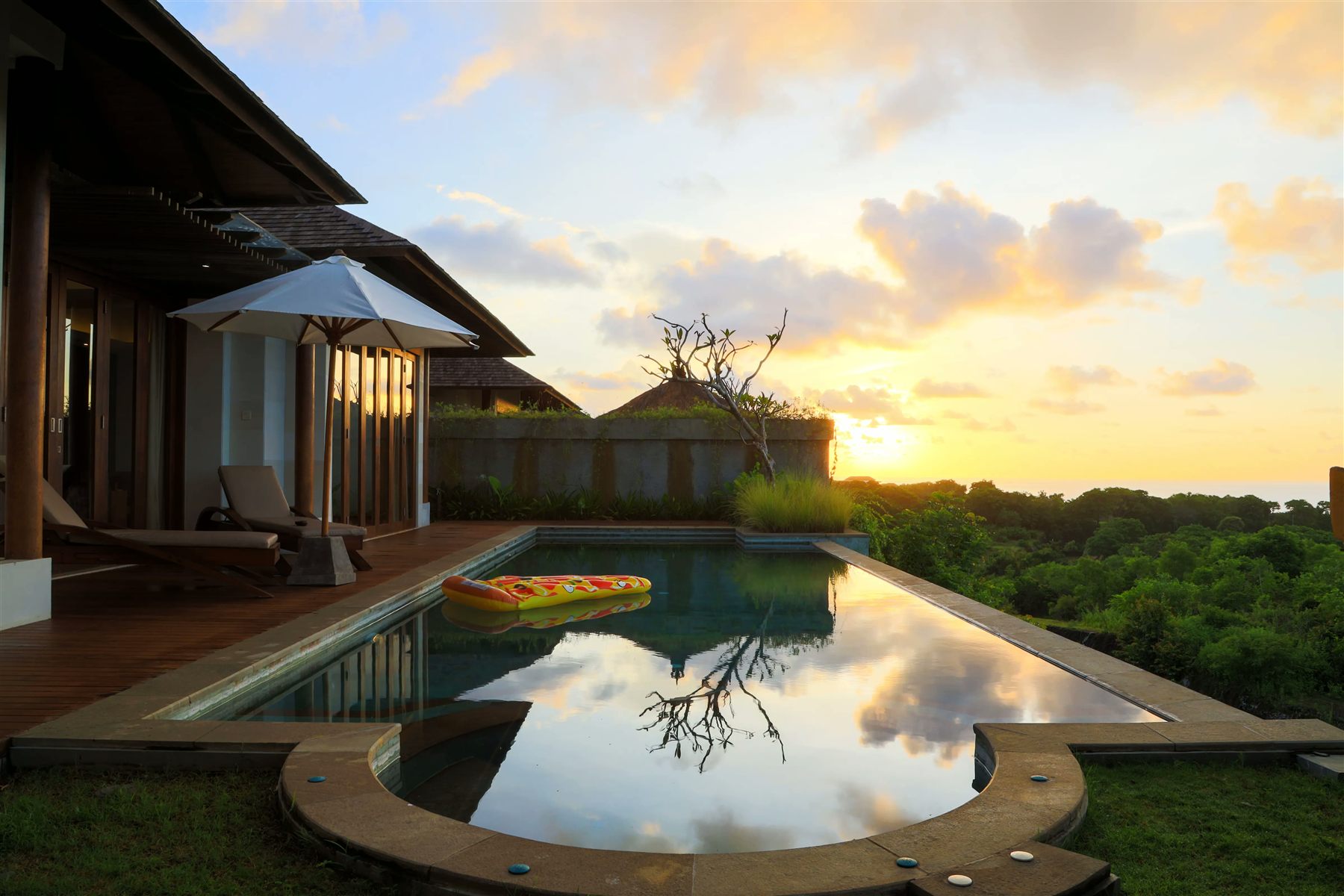 Pool at sunset reflecting a tree, beside a villa with a thatched roof