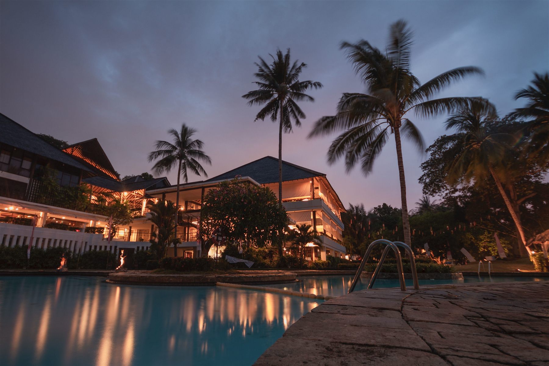 Pool at dusk in front of a lit villa with palm trees