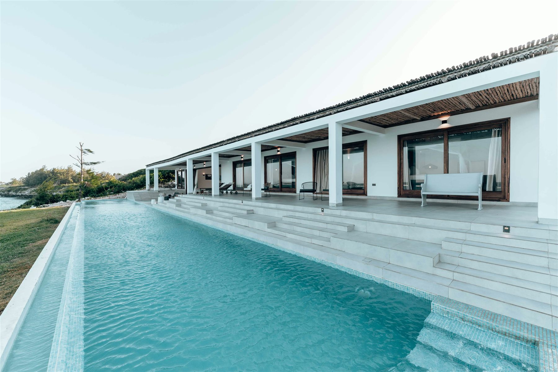 Long infinity pool beside a white single-storey villa at the coast, with open glass doors along the terrace